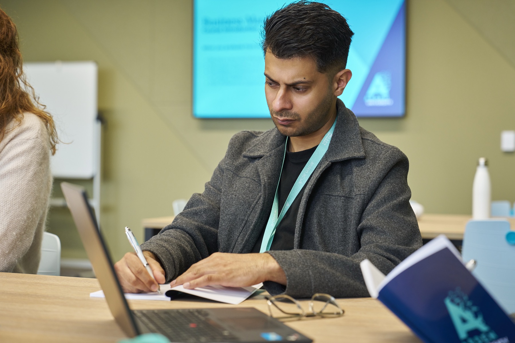 An Arden student making notes beside his laptop in a teaching room